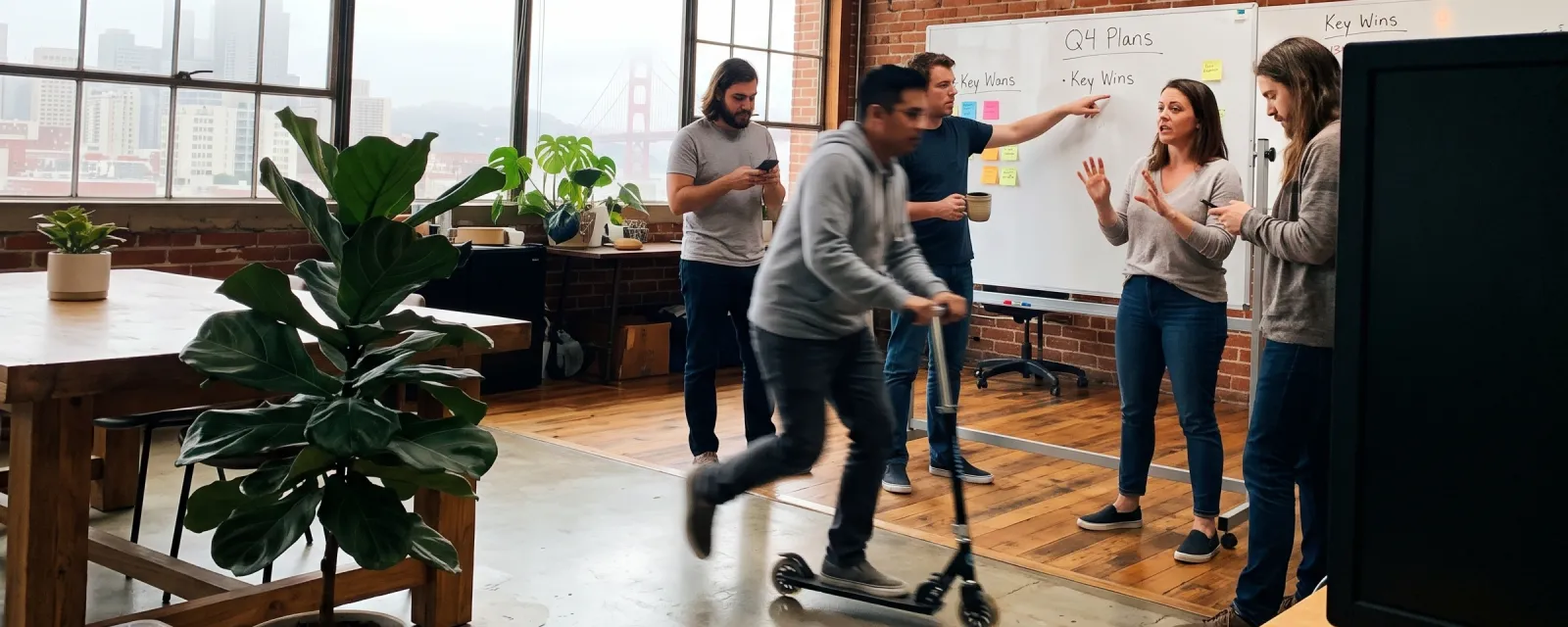 A teammate scooting through the Zeenly office mid-standup while others chat at a whiteboard, with the Golden Gate Bridge in the window behind